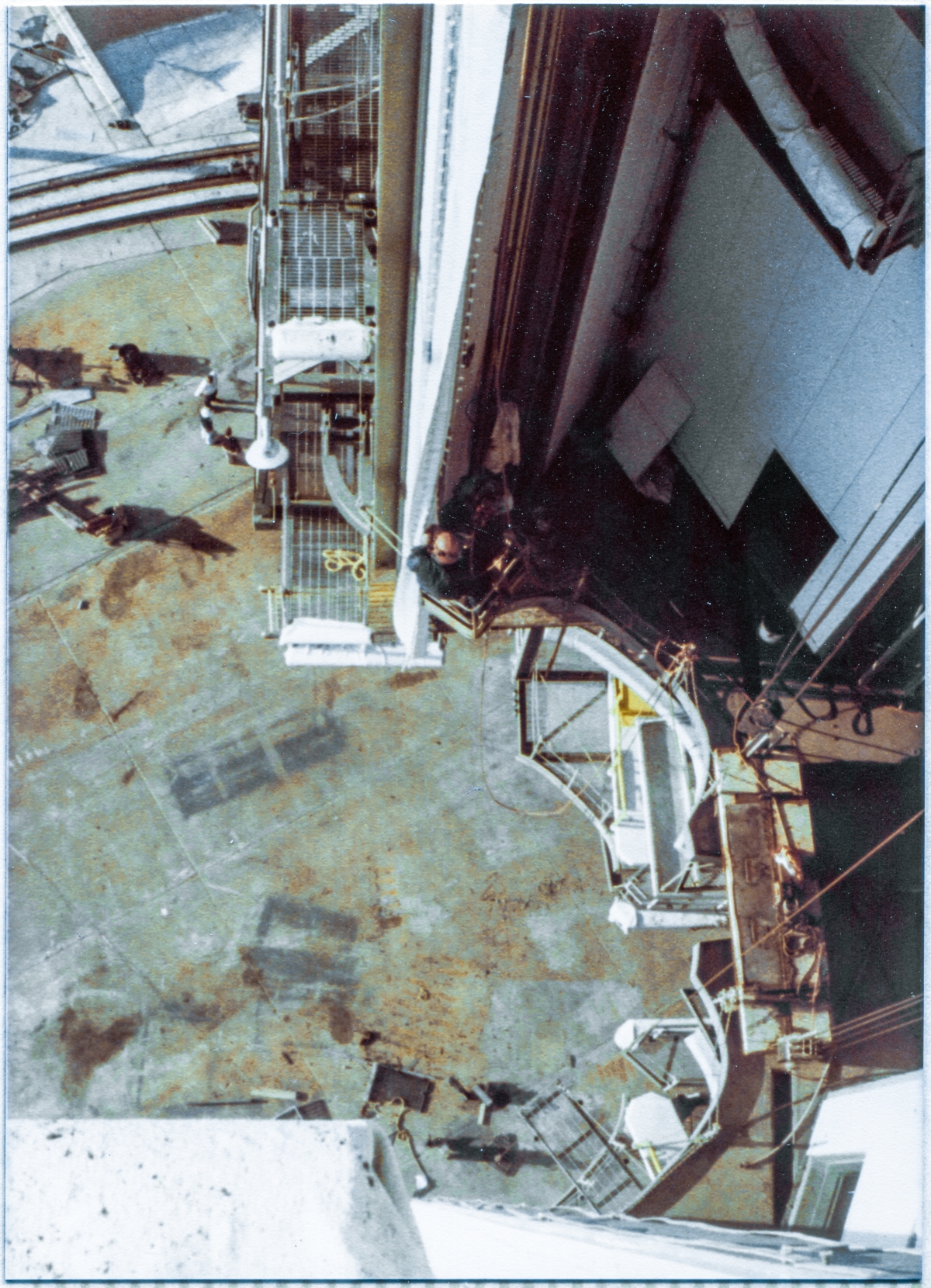 Image 038. Looking straight down, and across, from the Vehicle Access Platform at elevation  191’-0” on the Hinge Column side of the Rotating Service Structure at Space Shuttle Launch Complex 39-B, Kennedy Space Center, Florida. The concrete of the Pad Deck is 140 feet beneath you, and you can see a group of three people down there, in the upper left of the frame. To the right of them, just above the center of the frame, suspended in a spider basket, a Union Ironworker from Local 808, working for Wilhoit Steel Erectors, can be seen attaching a long white Inflatable Seal to the Right Orbiter Side Seal Panel. To the right of and below our ironworker, the distinctive shape of the Space Shuttle’s OMS Pods and Tail is outlined in the framing steel and platforms at elevations 135’-7” which is the Payload Changeout Room’s main floor level, 125’-0” which is the APU Servicing Platform level, and 112’-0” which is the APS Servicing Platform level, and is also the lowest main level of the RSS. When the RSS is swung around and mated to the Orbiter, the orbiter’s aft end will fill these distinctive shapes like a hand fitting inside a glove. Photo by James MacLaren.